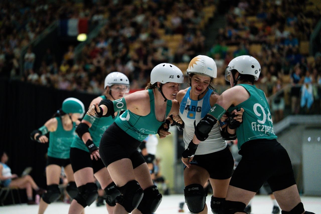 Ireland skaters in teal block an Argentina jammer in a tight pack, with a packed arena behind them.