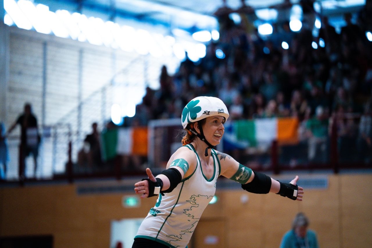 An Ireland skater in a shamrock helmet smiles with her arms spread wide, Irish flags in the stands behind her.