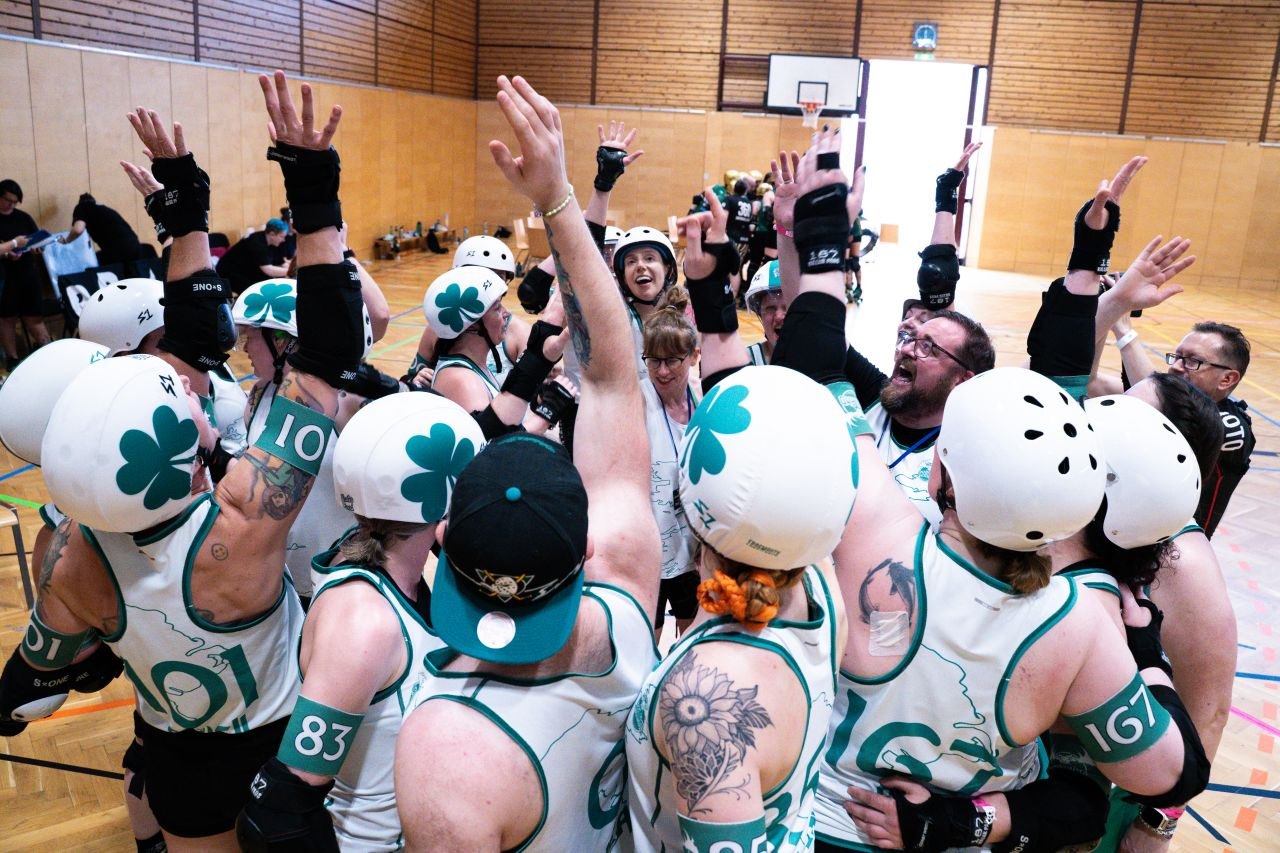 The Ireland team huddle with arms raised, cheering together in a sports hall.